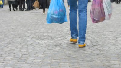 en erson går över ett torg. Hen bär flera kassar med mat. Vi ser bara hens ben,