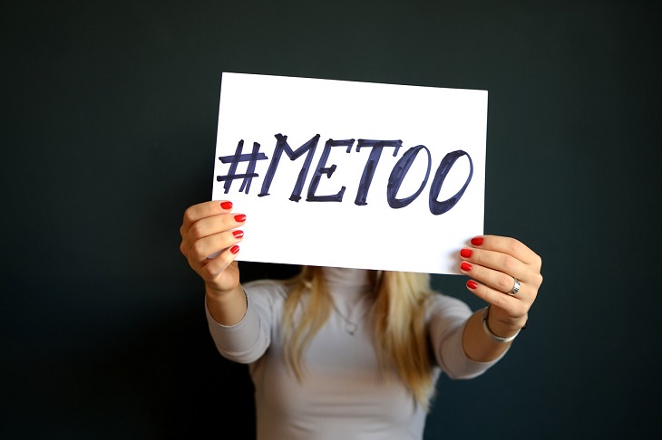 En tjej står mot en svart vägg. Vi ser inte hennes ansikte för framför sig sträcker hon fram en skylt där det står #metoo.