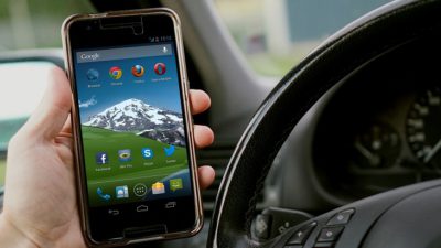 En hand håller i en mobil bredvid en ratt i en bil.