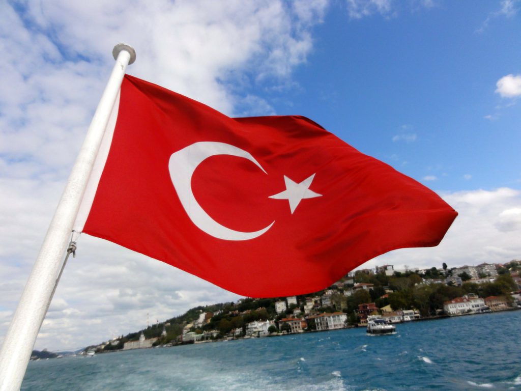 Turkiets flagga viftar i vinden över vatten. Det ser ut som att den sitter längst bak på en båt. Flaggan är röd med en vit halvmåne och en vit stjärna.