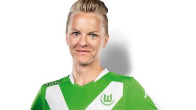 Nilla står mot en vit vägg och ler mot kameran. Hon har en grön fotbollströja och har kort hår som är bakåtkammat.