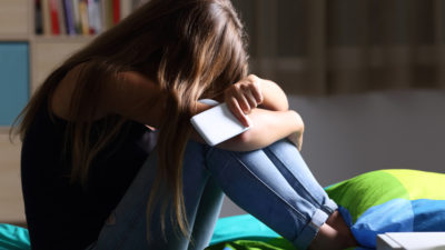 En tonåring sitter på en säng. Personen sitter med uppdragna knän, huvudet vilar mot knäna och den kramar om sina ben. I handen håller personen en telefon.