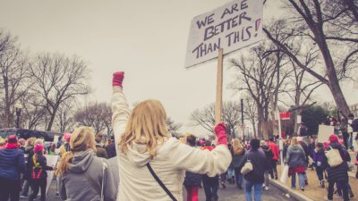 Bilden visar en manifestation för kvinnor. På bilden syns en person som håller i en skylt och knyter näven i luften. Runt om henne går personer iklädda rosa mössor. Mössorna är en symbol för tjejer och kvinnors rättigheter.